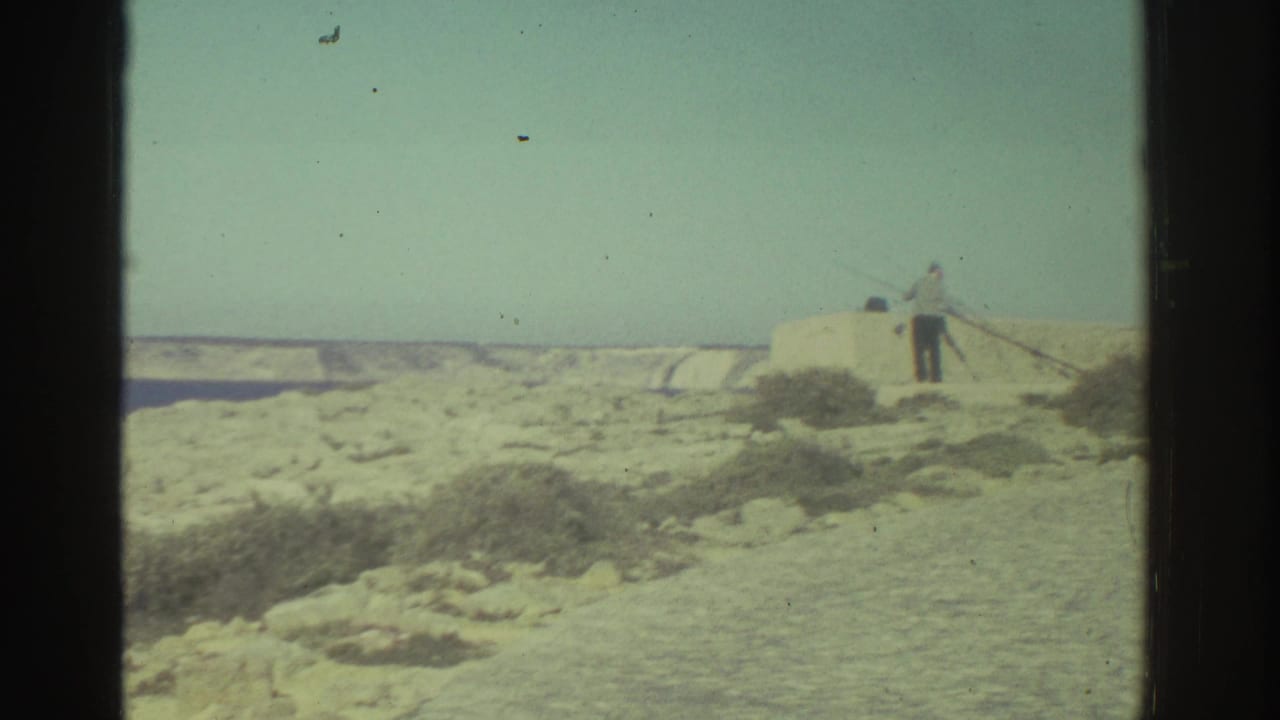 A man appears to fish from a pale stone wall on a rocky Greek coast, with blue water and distant land behind him.