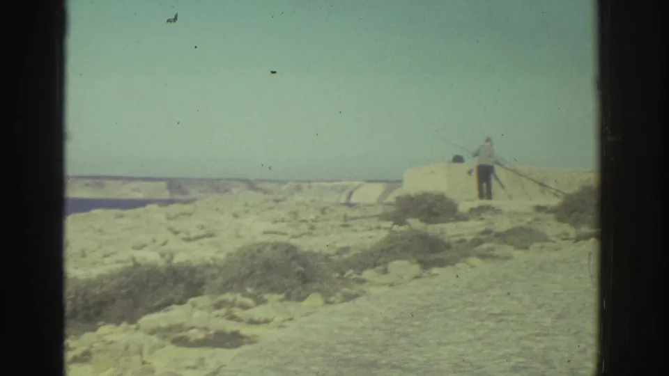 A man appears to fish from a pale stone wall on a rocky Greek coast, with blue water and distant land behind him.