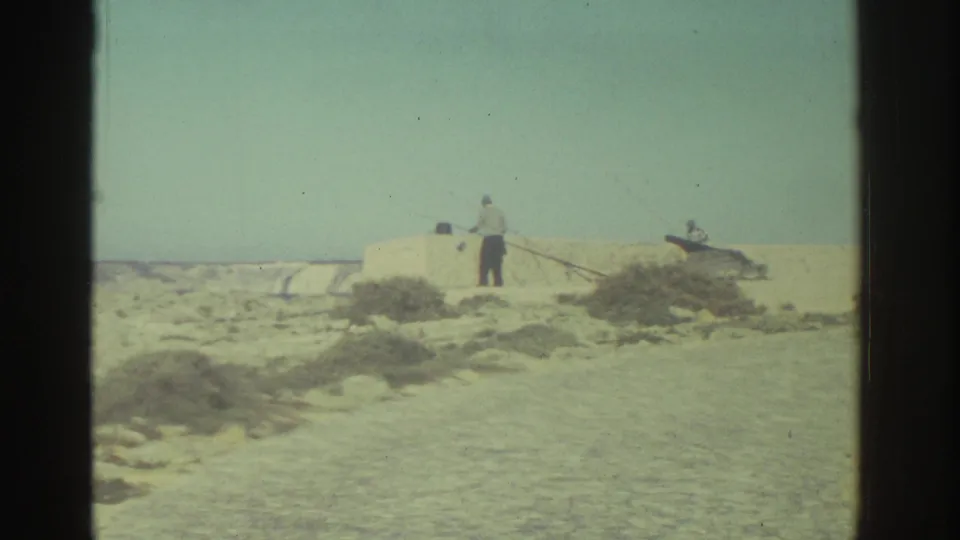 The distant figure stands with a fishing rod beside a low wall on an arid coastal path in Greece, with the sea beyond.