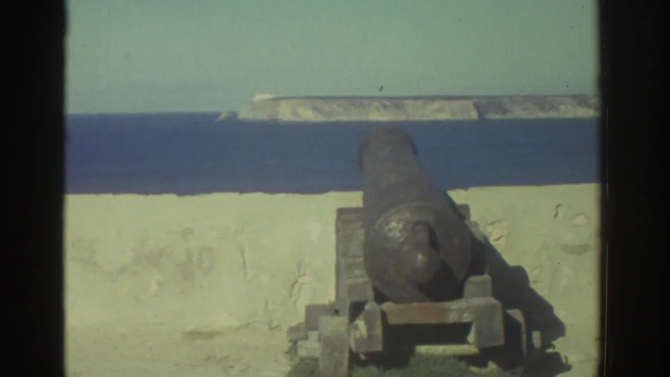 A weathered cannon sits on a seaside wall in Greece, aimed toward blue water and a pale headland in 1976 8mm footage.