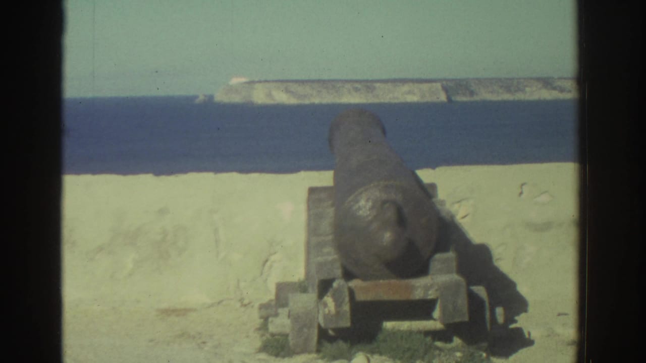 1976 Greece 8mm frame showing a rusted coastal cannon on a mount beside a pale wall, with blue water and rocky shoreline in the distance.