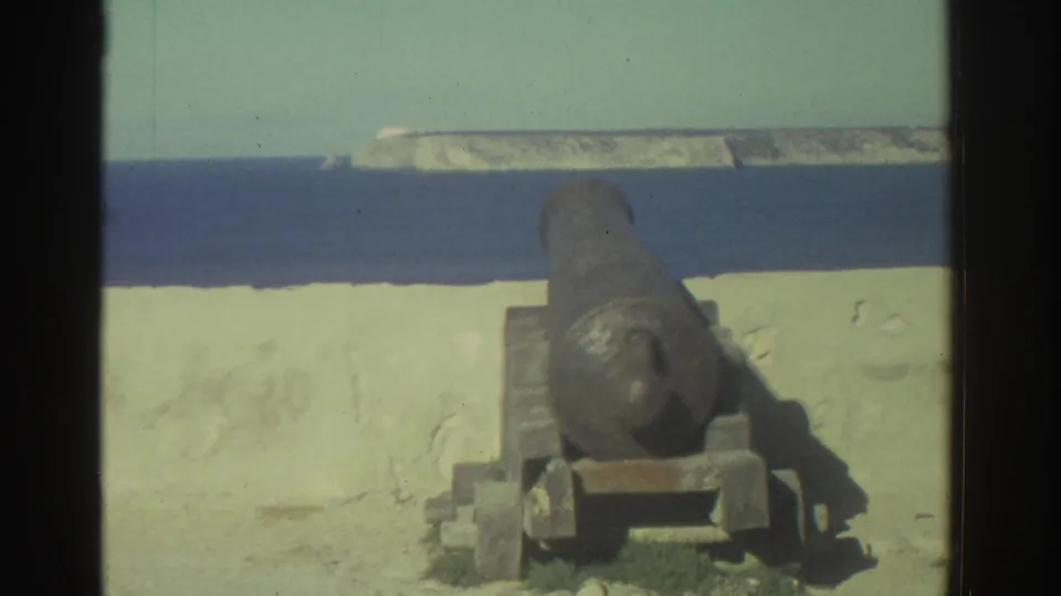 1976 Greece 8mm frame showing a rusted coastal cannon on a mount beside a pale wall, with blue water and rocky shoreline in the distance.