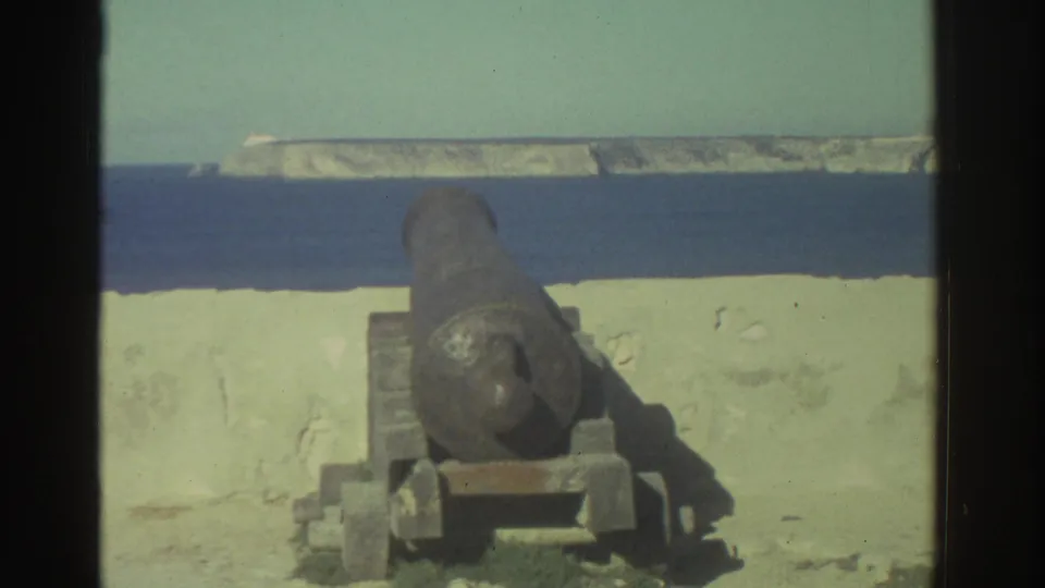 A rusted cannon at a Greek coastal fortification is framed against a pale wall, blue water, and a distant rocky coastline.