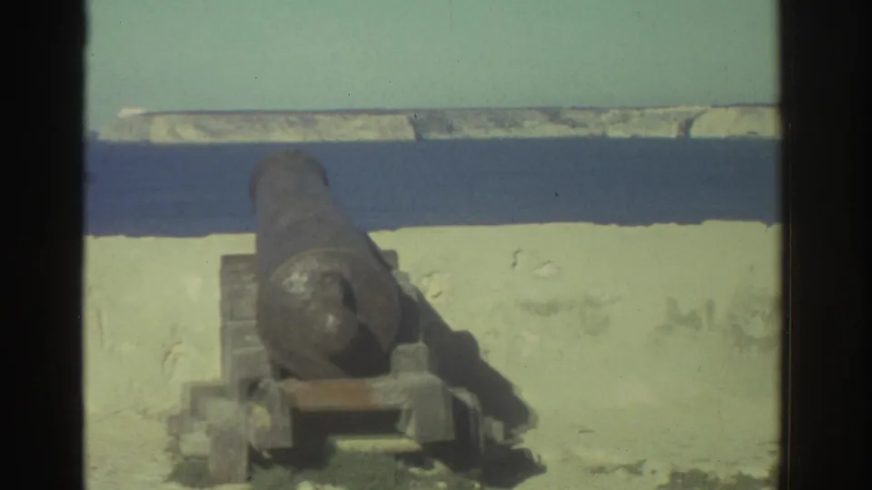 Close 8mm view of a weathered cannon and mount at a Greek seaside fortification, with pale masonry and blue water behind.