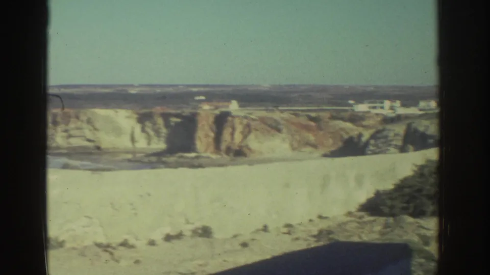 The frame opens to a wider Greek coastal view, showing a pale fort wall, cliffs, blue sea, and small distant buildings.