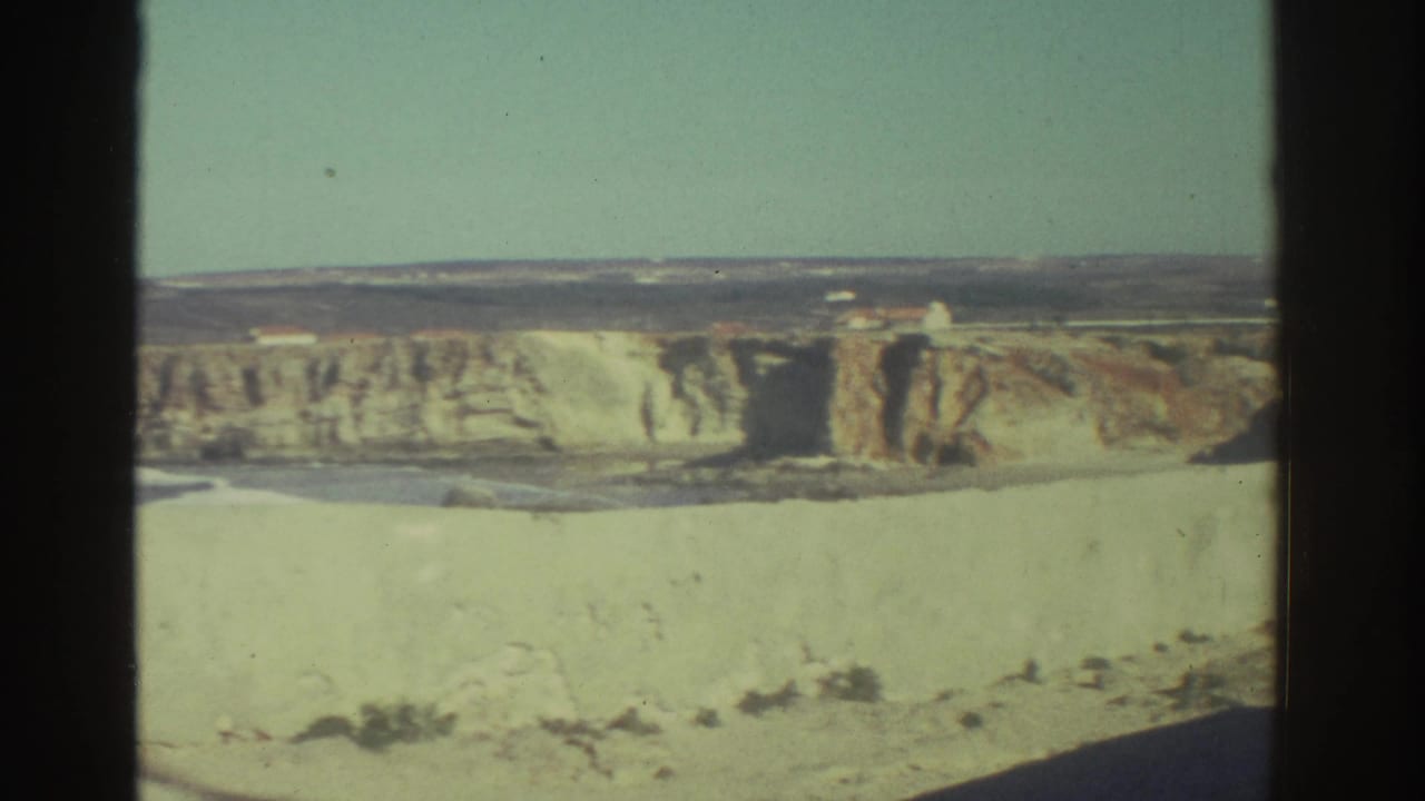1976 Greece 8mm frame showing a distant pale cliff or fortified wall, open sky, and small structures along the horizon.