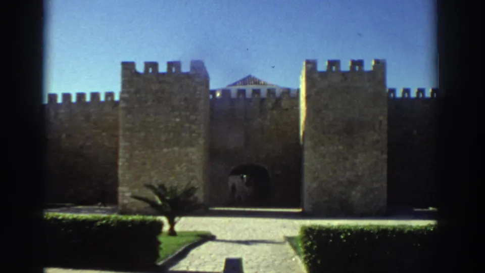 1976 Greece 8mm frame showing a crenellated stone castle exterior, central arched gateway, trimmed hedges, path, topiary, and a distant person.