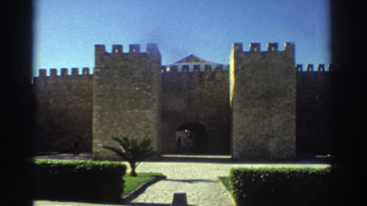 1976 Greece 8mm frame showing a stone fortress gate with square towers, a paved approach, trimmed hedges, grass, and a small palm tree.