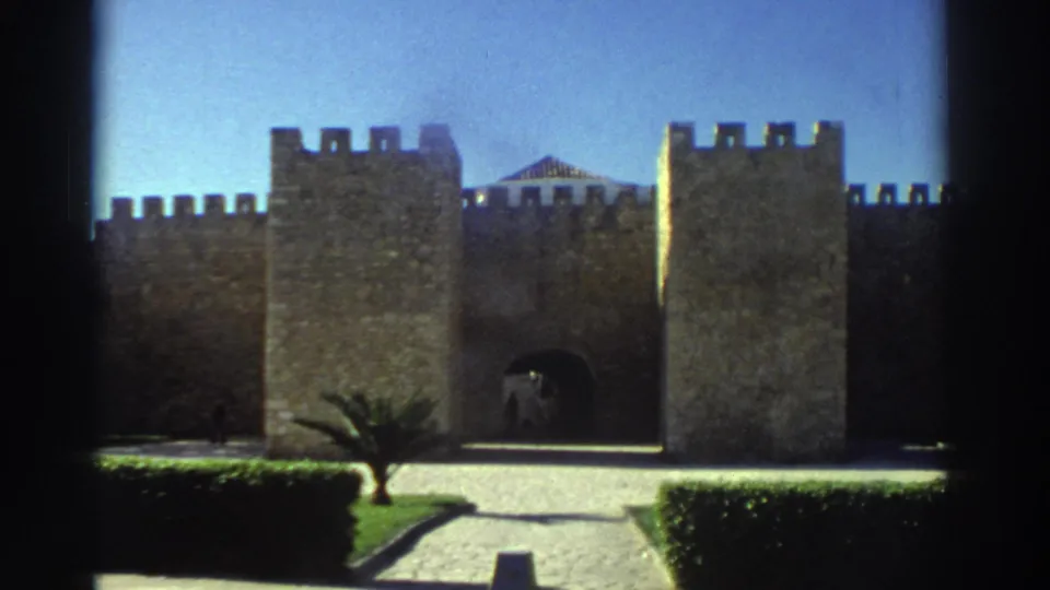 1976 Greece 8mm frame showing a stone fortress gate with square towers, a paved approach, trimmed hedges, grass, and a small palm tree.
