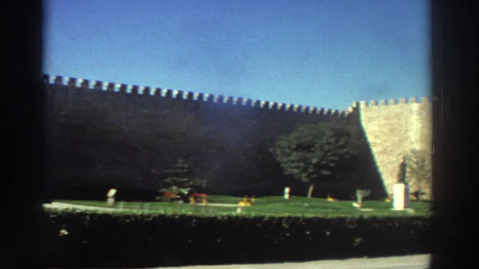 1976 Greece 8mm frame showing the fortress wall continuing toward a bright corner tower, with lawn, shrubs, a path, and a pedestal statue.