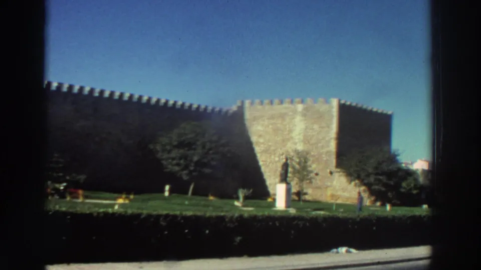 1976 Greece 8mm frame of a fortress corner beside landscaped grass, trees, and a pedestal statue, framed by dark scan borders.