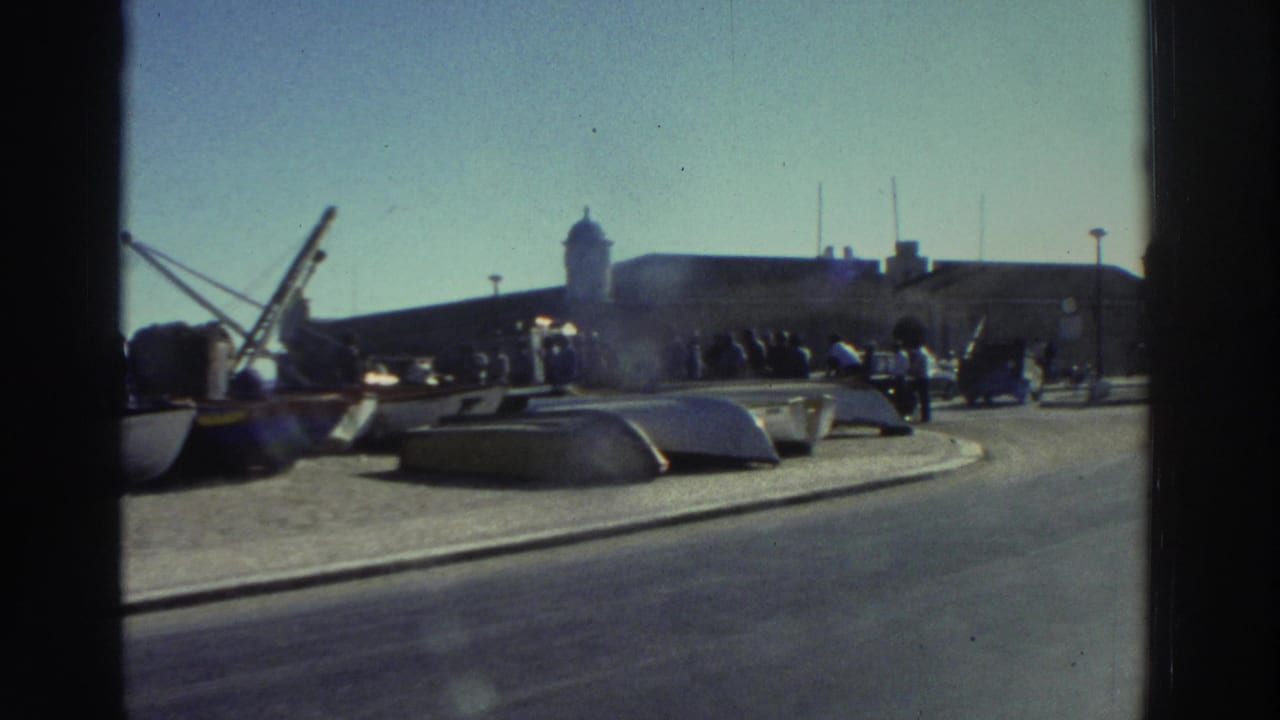 1976 Greece harbor view with small boats on a paved quay, a crane, distant people, roadway, and a domed waterfront building.