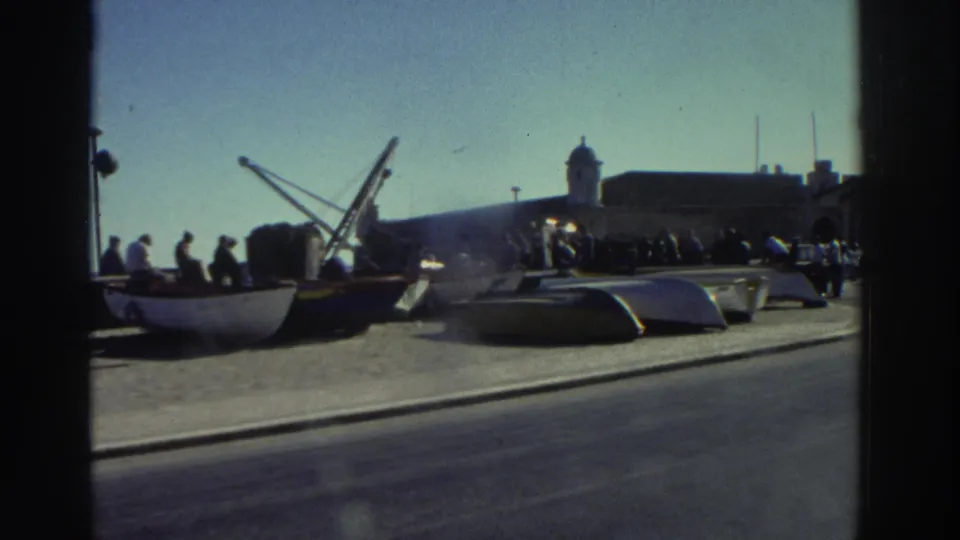 1976 Greece harbor frame showing people in casual clothing standing and sitting near a small white boat on the quay.