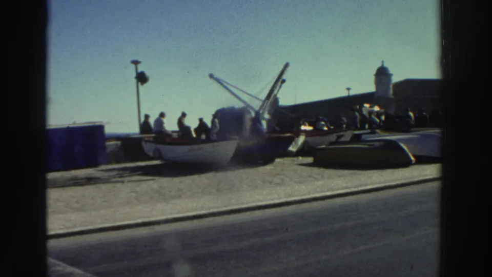 1976 Greece home-movie frame with boats on the quay, people standing nearby, a crane, roadway, and distant harbor architecture.