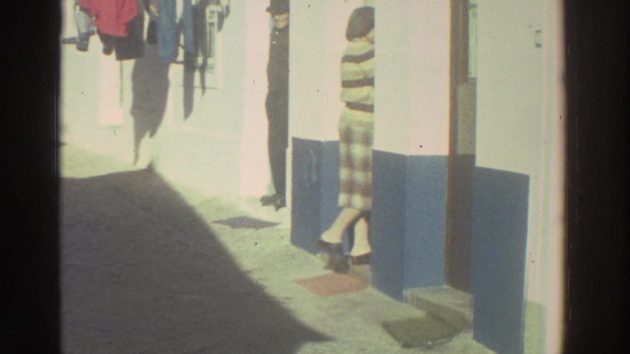 1976 Greece home-movie frame showing a bright narrow residential alley with laundry outside and a person standing near a doorway.