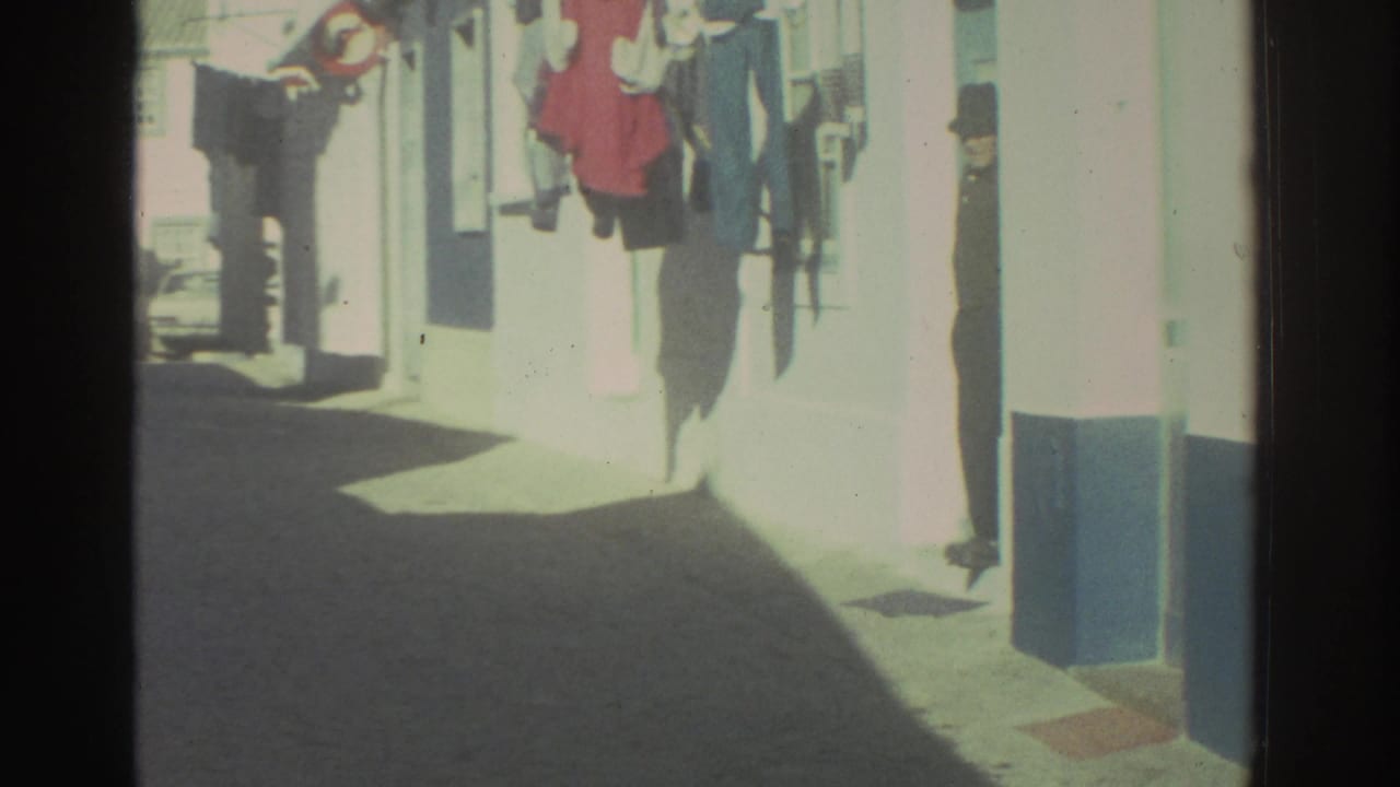 1976 Greece street frame with whitewashed buildings, laundry lines, a narrow lane, and a distant person by an entrance.
