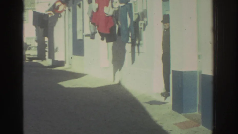 1976 Greece street frame with whitewashed buildings, laundry lines, a narrow lane, and a distant person by an entrance.