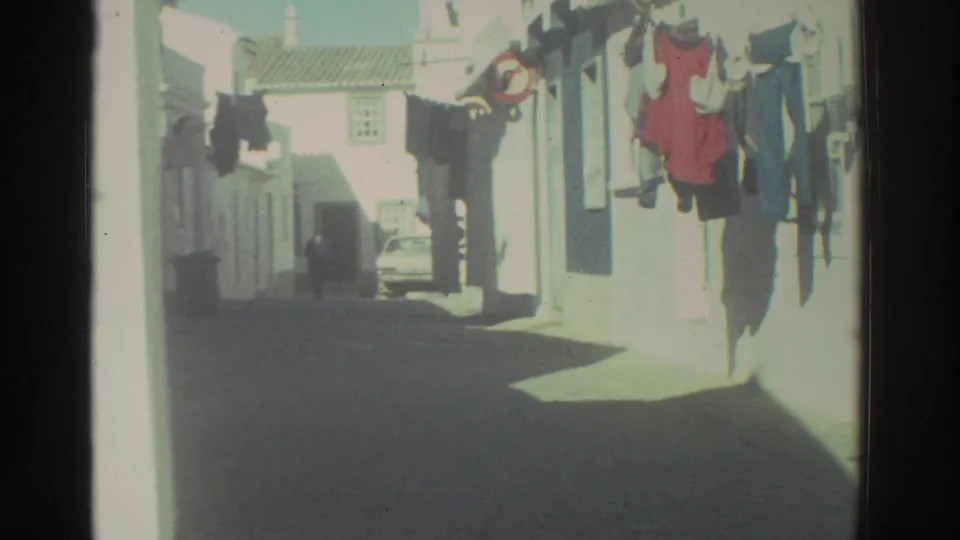 1976 Greece home-movie view down a narrow residential street with laundry on the right and a small car farther back.
