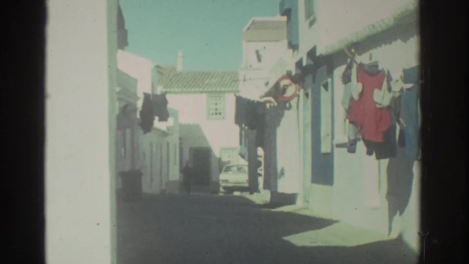 1976 Greece frame looking along a residential alley with hanging clothes, white walls, tiled roofs, and a car in the distance.