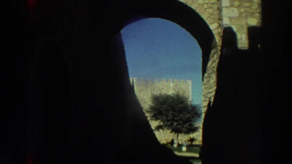 1976 Greece 8mm frame looking from a dark stone arch toward a bright castle wall, tree, and clear blue sky.
