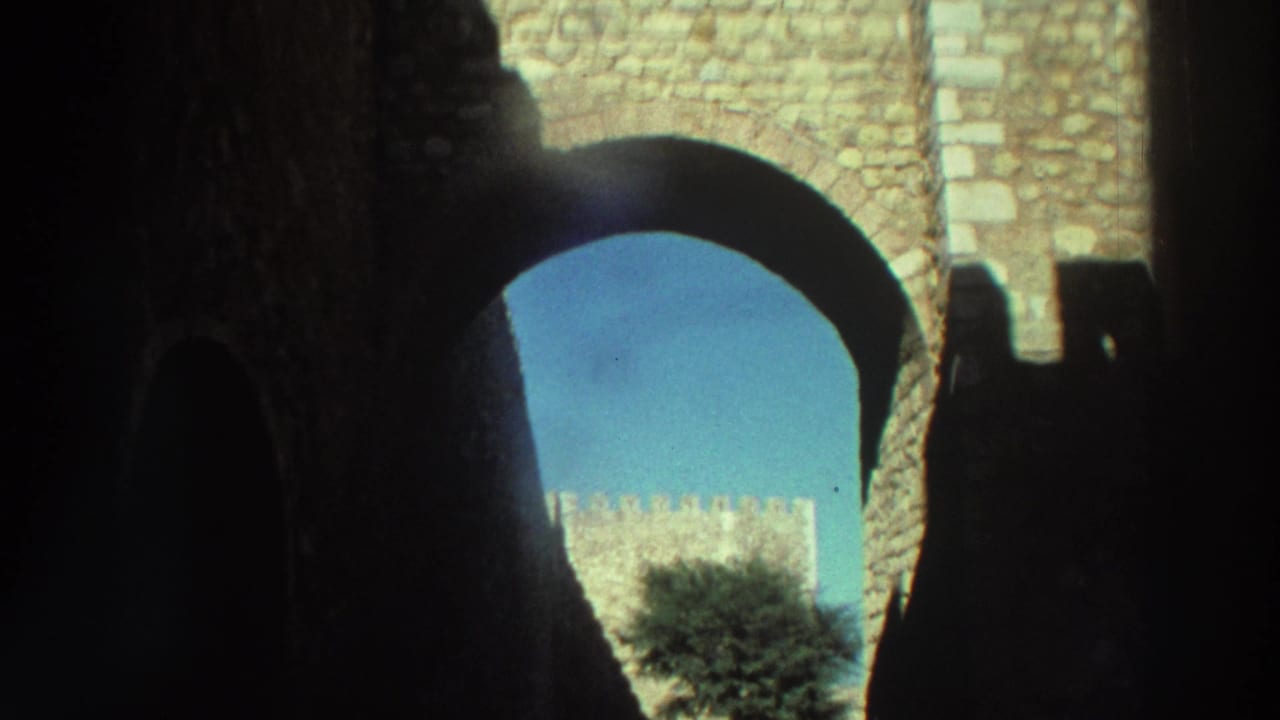 Handheld 8mm view from within a shadowed Greek castle passage toward bright masonry, sky, and courtyard greenery.