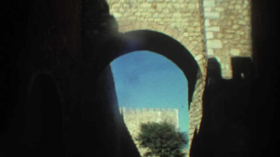 Handheld 8mm view from within a shadowed Greek castle passage toward bright masonry, sky, and courtyard greenery.