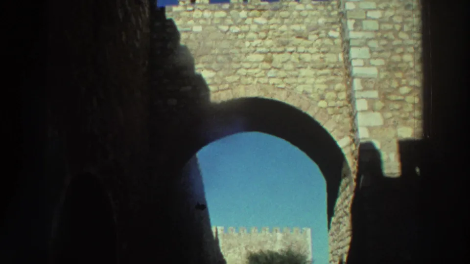 1976 Greece 8mm frame of a castle arch in heavy shade, with sunlit stonework, sky, and a tree visible through the opening.