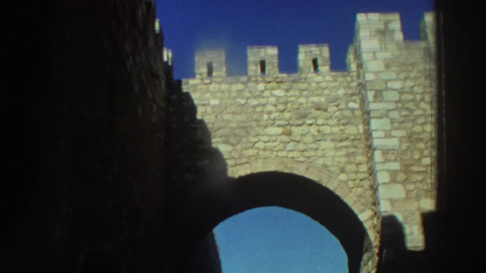 Color 8mm frame from Greece showing crenellated castle masonry rising over a shaded arch beneath a vivid blue sky.