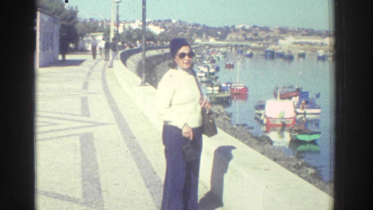 Woman in sunglasses stands on a harbor promenade with small boats behind her.