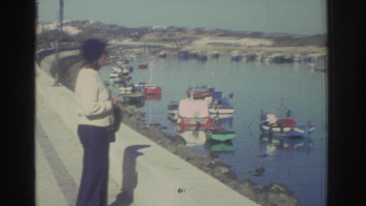 A woman in 1976 Greece faces the harbor from a concrete promenade, with colorful small boats and a low shoreline visible across the water.