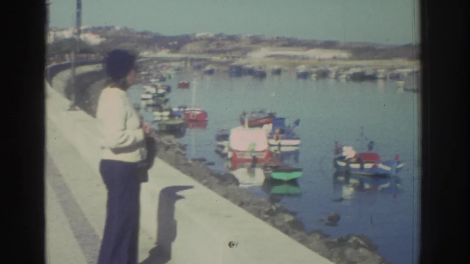 A woman in 1976 Greece faces the harbor from a concrete promenade, with colorful small boats and a low shoreline visible across the water.
