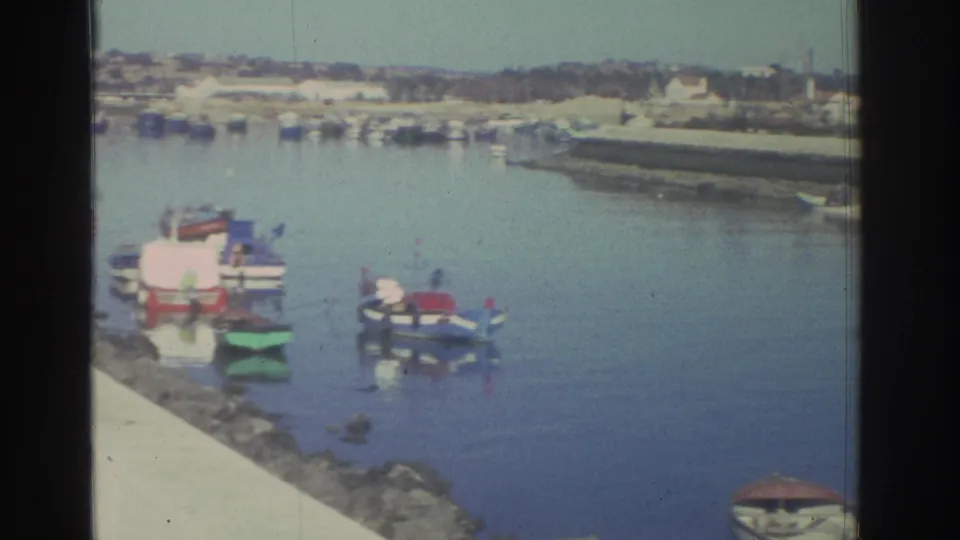 A 1976 Greece harbor view shows small boats on still blue water beside rocks and a concrete promenade edge, with shoreline beyond.