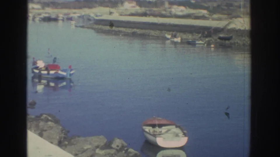 A quiet Greek harbor scene from 1976 shows small boats, calm water, rocks in the foreground, and a low waterfront wall in the distance.