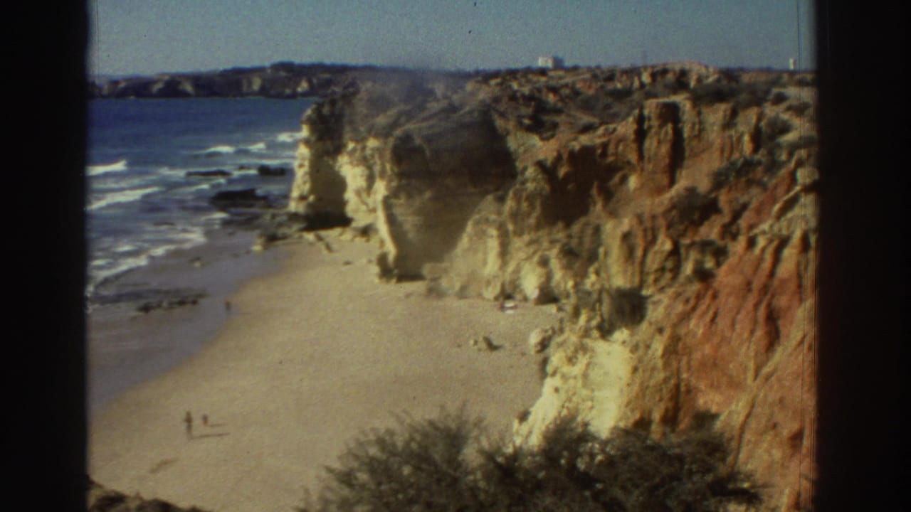 1976 Greece 8mm frame showing a sandy beach beneath ochre cliffs, with blue sea, surf lines, and small distant beach figures.