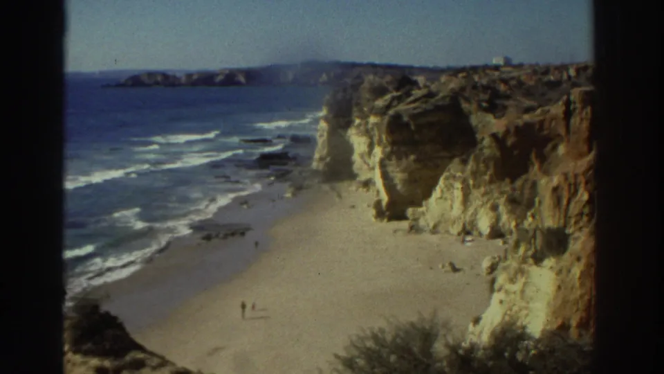 1976 Greece home-movie frame of a beach tucked below pale and ochre cliffs, with gentle surf and distant people walking on the sand.