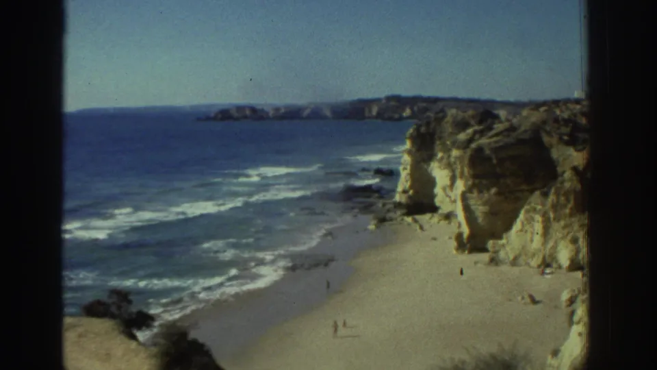 1976 Greece 8mm frame showing a broad view of surf, sand, layered cliff walls, and tiny beach figures from an elevated position.