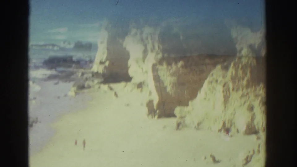 Soft, bright 1976 Greece 8mm frame of pale coastal cliffs and beach, with the sea beyond and distant people visible near the sand.