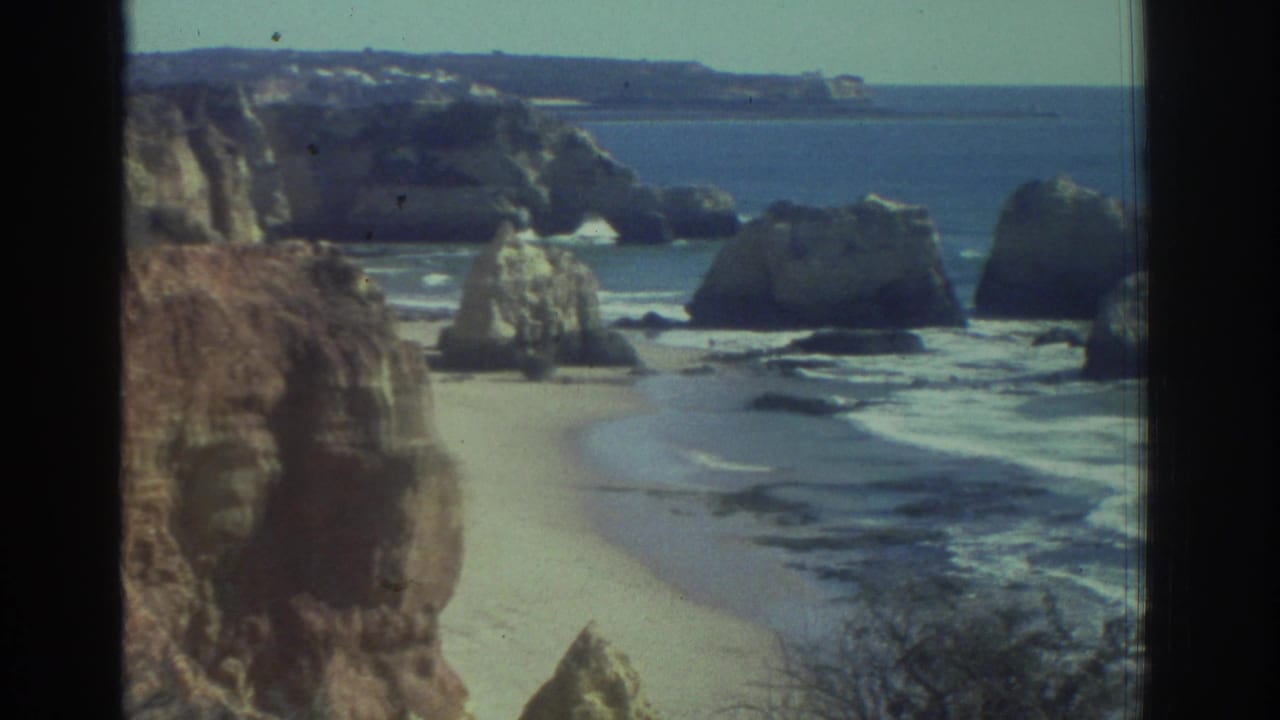 1976 Greece 8mm frame showing a rugged beach, pale sand, cliff faces, offshore rock formations, blue water, and dark frame edges.