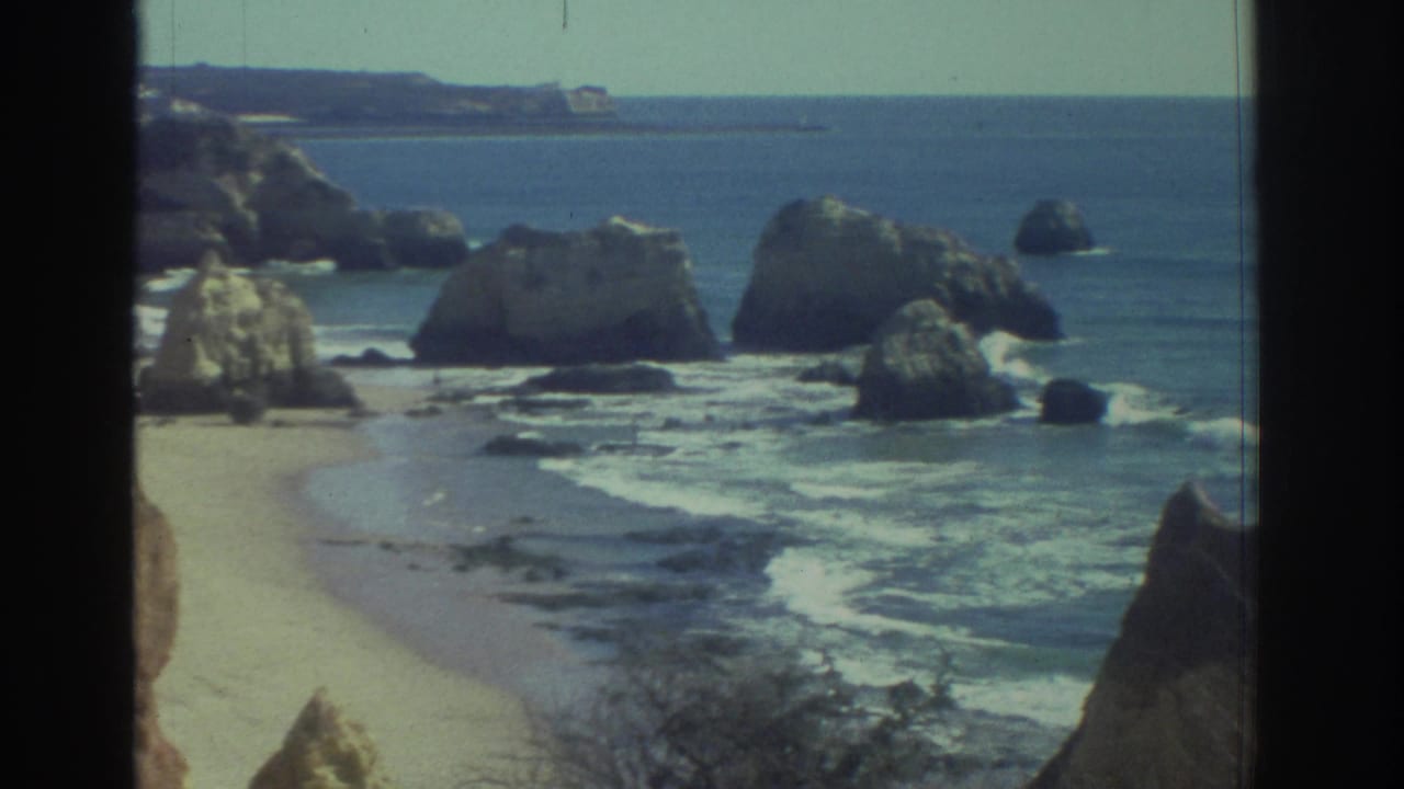 1976 Greece 8mm view of a rocky shoreline, sandy cove, scattered sea rocks, gentle surf, and blue water.