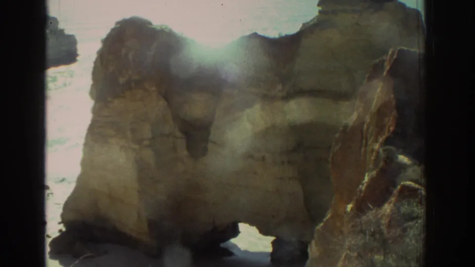 1976 Greece 8mm frame with a closer view of weathered coastal rocks, strong sun glare, pale surf, and visible frame borders.