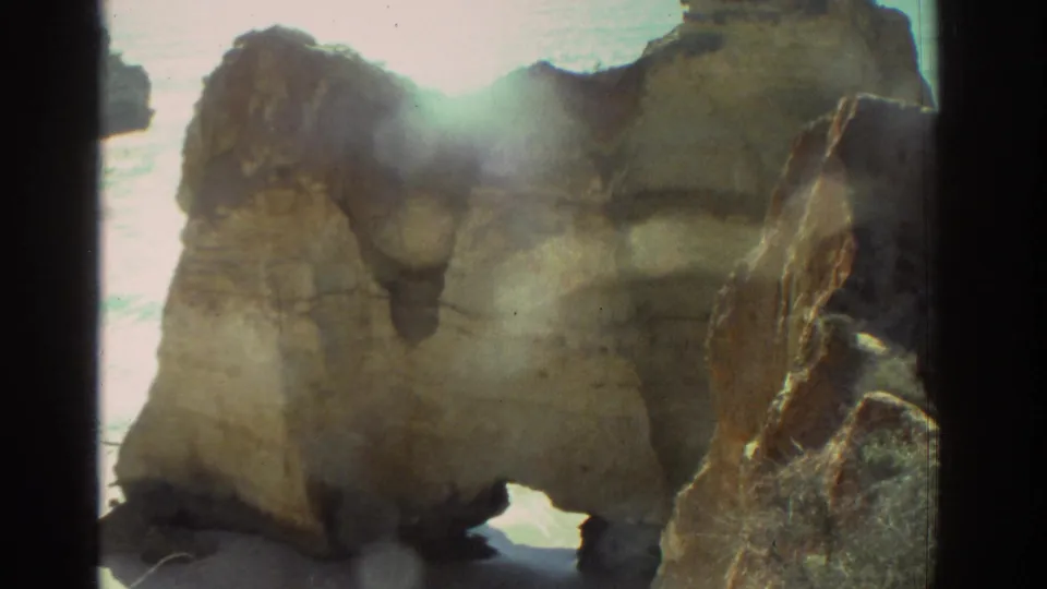 1976 Greece 8mm frame showing weathered rocks near the shoreline, bright reflected sea light, soft focus, and film-frame borders.