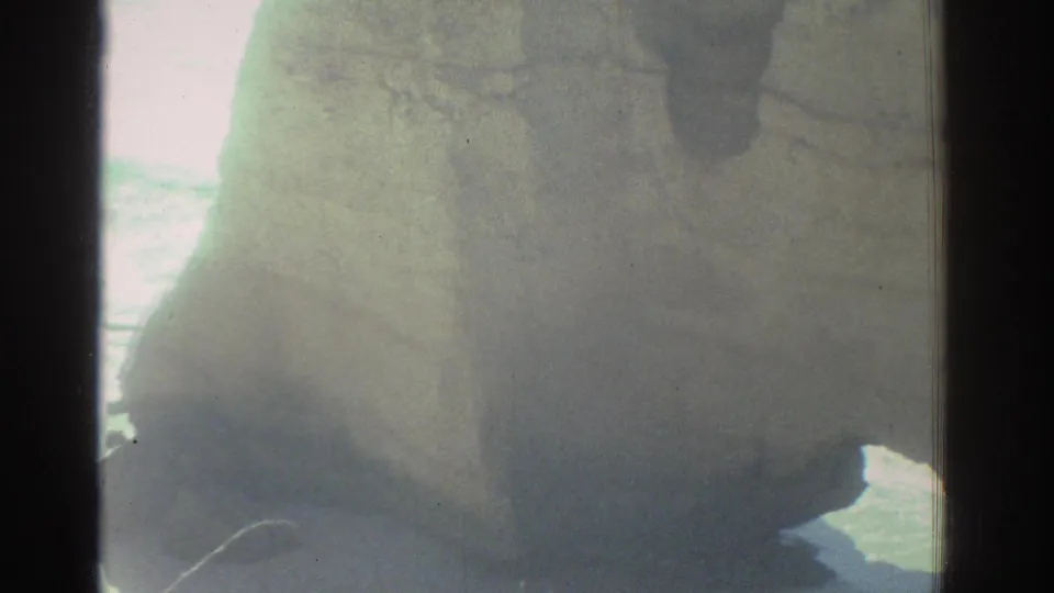 A soft, flared 8mm view of a large Greek coastal rock face with bright sea light behind it.