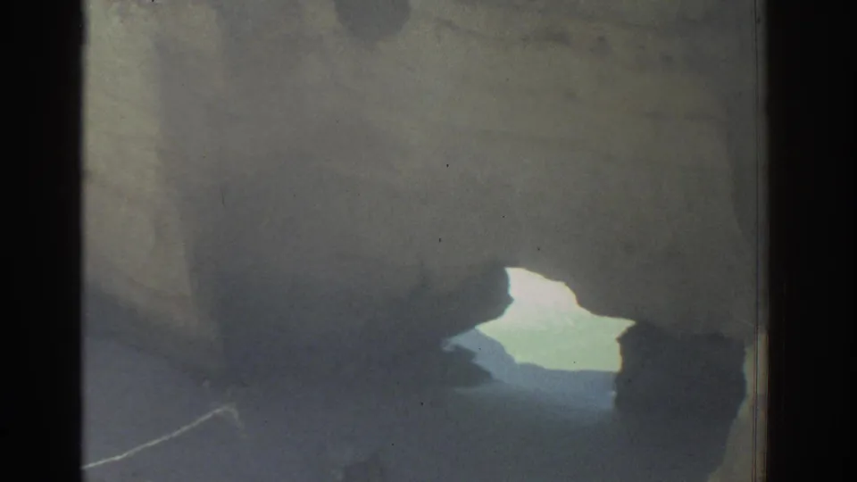 Shadowed coastal rocks in Greece, filmed on 8mm, with a small bright arch opening toward the water.
