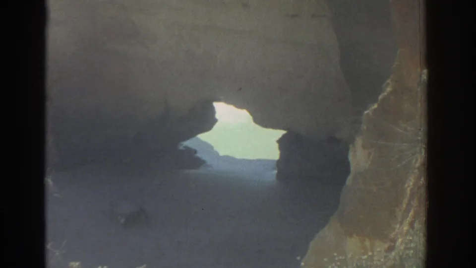 A 1976 Greece home-movie frame looking through a coastal rock opening toward bright sea light.