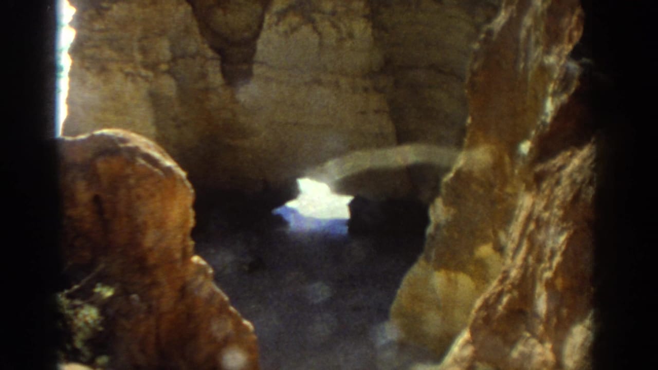 Handheld 8mm view of warm-toned Greek coastal rocks framing a small bright opening toward the beach or sea.
