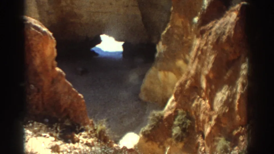 1976 Greece home-movie frame moving through textured coastal rock toward a small sunlit opening beyond.