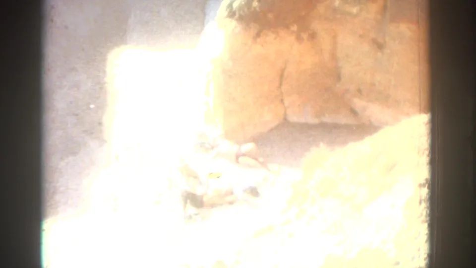 Bright 1976 Greece beach frame showing two distant sunbathers on sand beside a warm-toned rock formation.