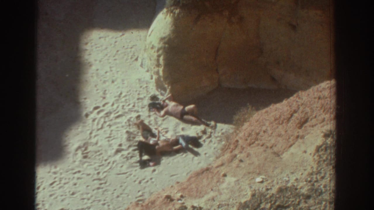 Two beachgoers rest in bright sun near large rock formations in 1976 Greece home-movie footage.