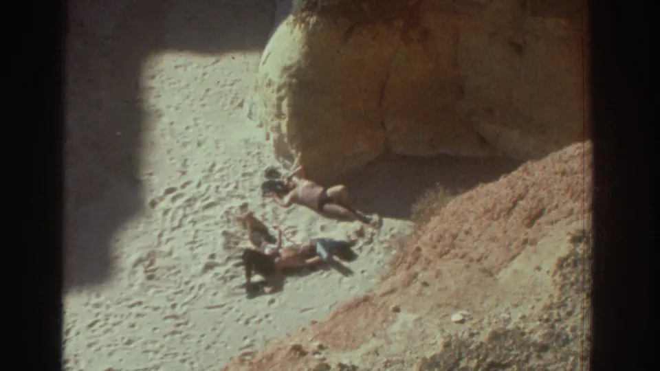 Two beachgoers rest in bright sun near large rock formations in 1976 Greece home-movie footage.