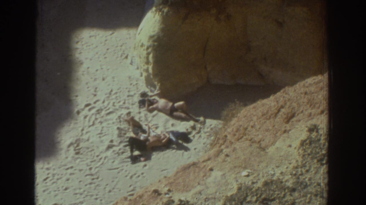 Beachgoers lie near a rock face on a bright Greek coastal beach in 1970s 8mm footage.